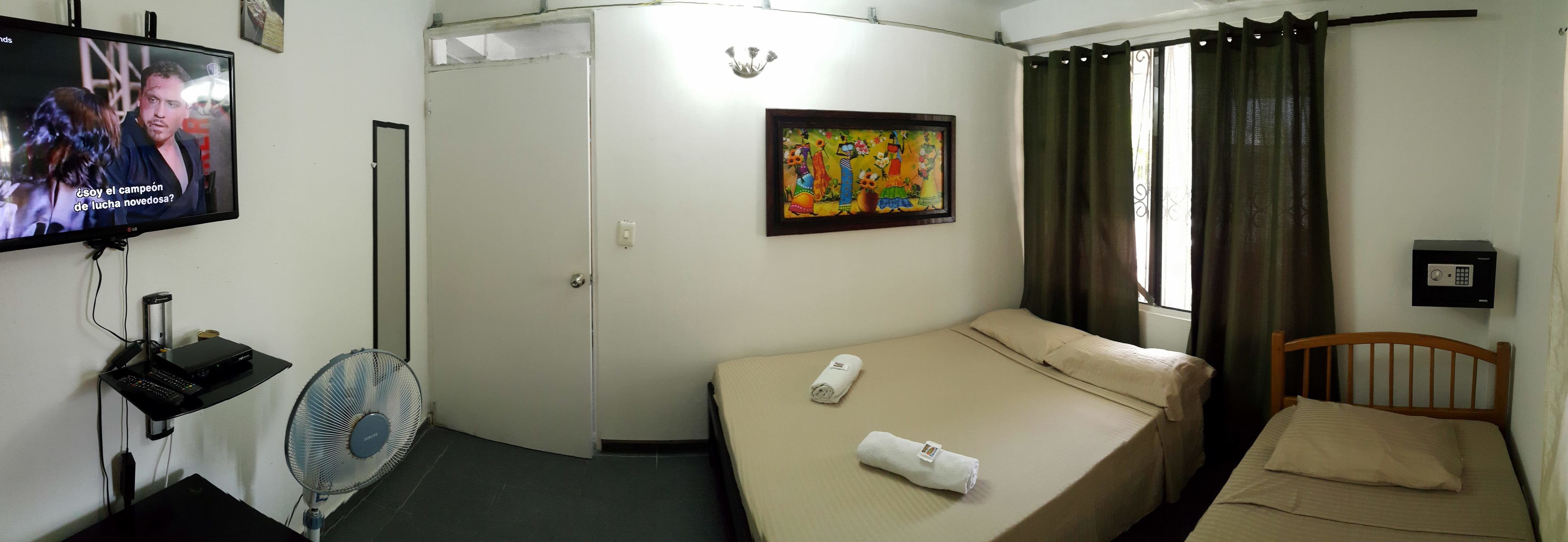 Una habitación de hotel con una cama, televisión, ventilador, un cuadro y una ventana con cortinas.