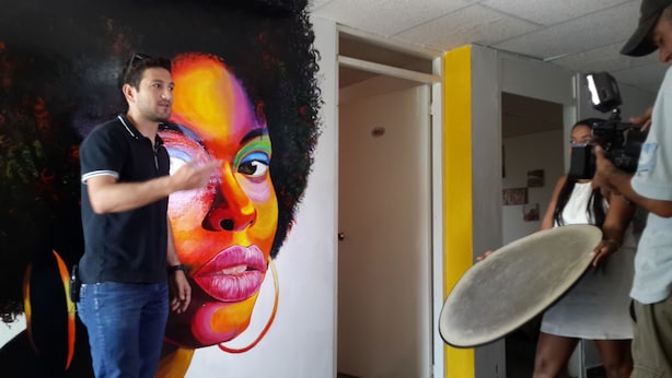 Une personne se tient devant une fresque murale aux couleurs vives.