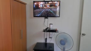 Un téléviseur fixé au mur diffusant un concert, une petite étagère avec des appareils électroniques et un ventilateur sur pied.