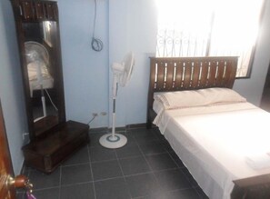 A bedroom with a bed, a standing fan, a wooden wardrobe, and a window with bars.