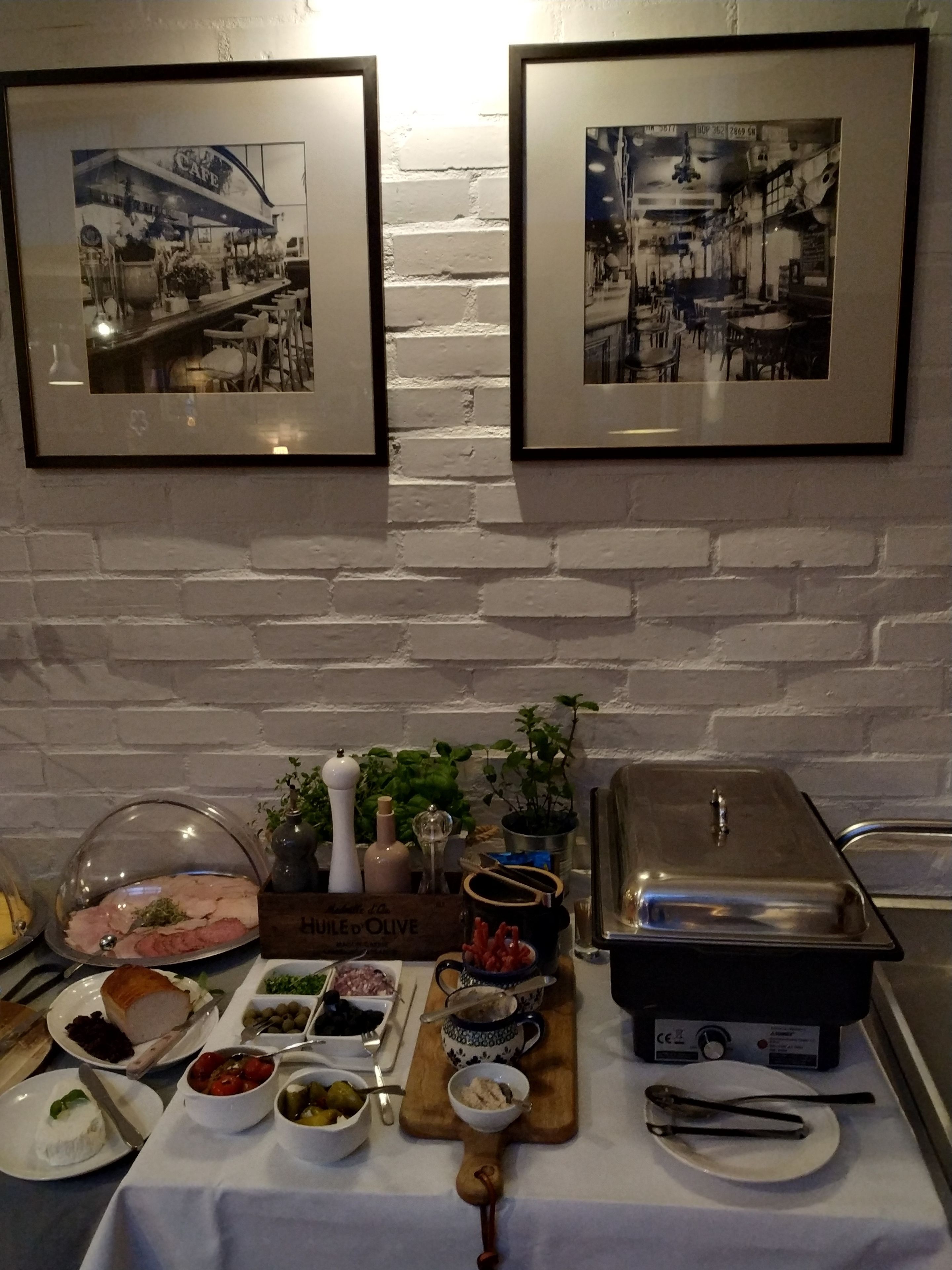 Une table de buffet garnie de divers plats et équipée d’un appareil pour maintenir les aliments au chaud, le tout placé devant un mur de briques blanches, avec deux tableaux encadrés accrochés au-dessus.