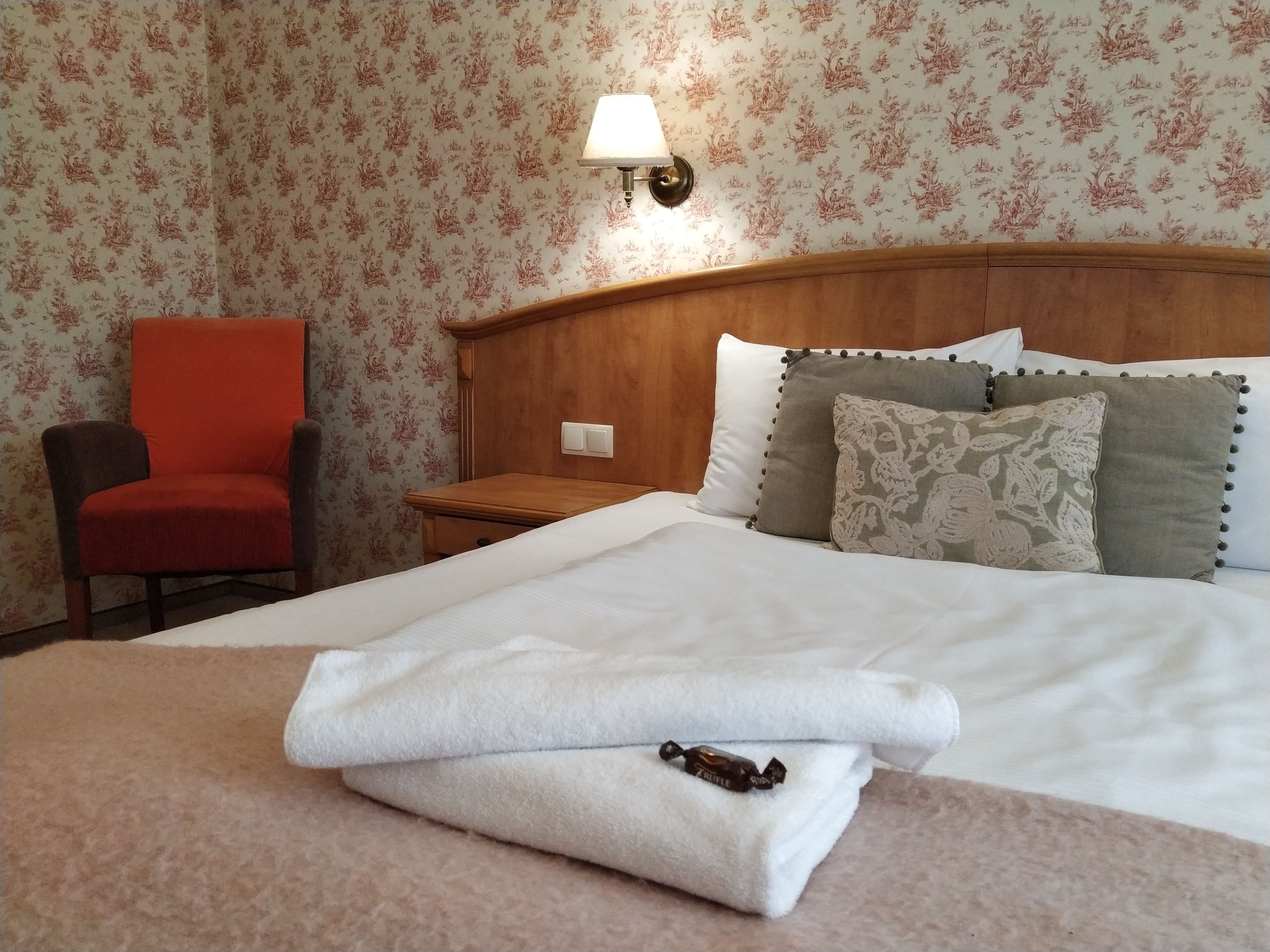 A neatly made bed with white linens, a red chair, and a wooden nightstand with a lamp.