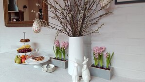 Een eettafel met een presentatie van fruit en gebak, een vaas met takken en bloemen, en twee witte konijnenbeeldjes.