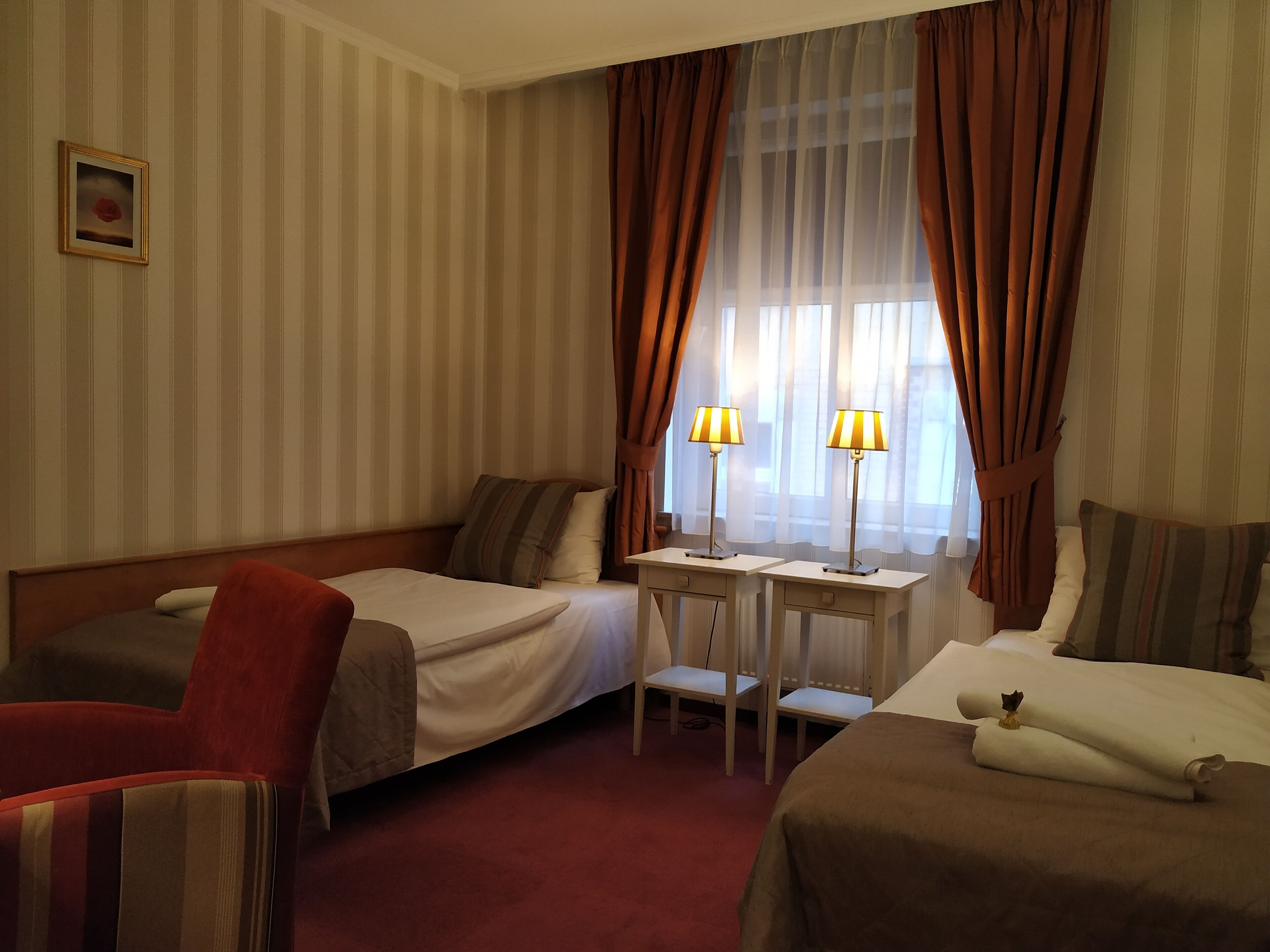 A hotel room with two beds, a small table with lamps, a chair, and a window with curtains.