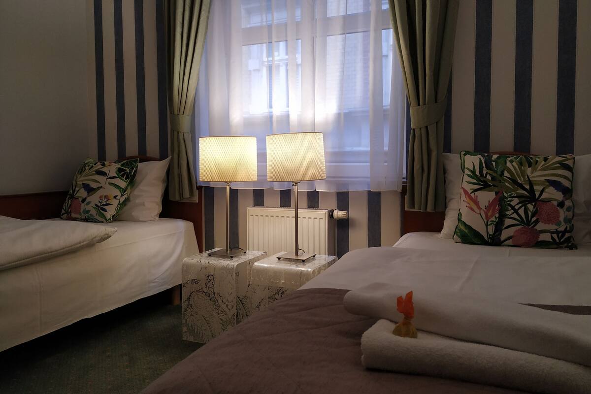 Een hotelkamer met twee bedden, gestreepte gordijnen, een radiator en een klein tafeltje met lampen.