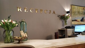 Een receptiebalie met een marmeren blad, een houten onderkast met sierlijke handgrepen, een bordje aan de muur met de tekst 'RECEPCJA', een computermonitor, een telefoon en decoratieve objecten waaronder een vaas met bloemen en een glazen fles met papieren.