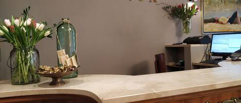 A reception desk with a marble top, a wooden base with ornate handles, a wall-mounted sign reading "RECEPCJA", a computer monitor, a telephone, and decorative items including a vase with flowers and a glass bottle with papers.