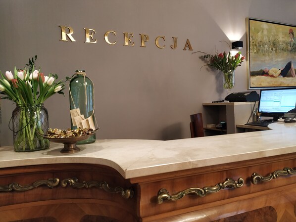 A reception desk with a marble top, a wooden base with ornate handles, a wall-mounted sign reading "RECEPCJA", a computer monitor, a telephone, and decorative items including a vase with flowers and a glass bottle with papers.