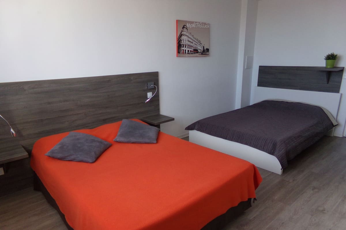 Une chambre avec deux lits, l’un recouvert d’une couverture orange et l’autre d’une couverture foncée. On y trouve une petite étagère sur laquelle est posée une plante, ainsi qu’un tableau encadré accroché au mur.