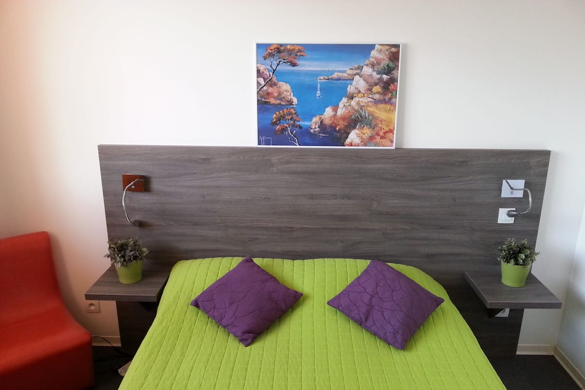Une chambre d’hôtel avec une couvre-lit verte, deux coussins violets, une chaise rouge, deux petites plantes en pot et un tableau représentant un paysage côtier accroché au mur.