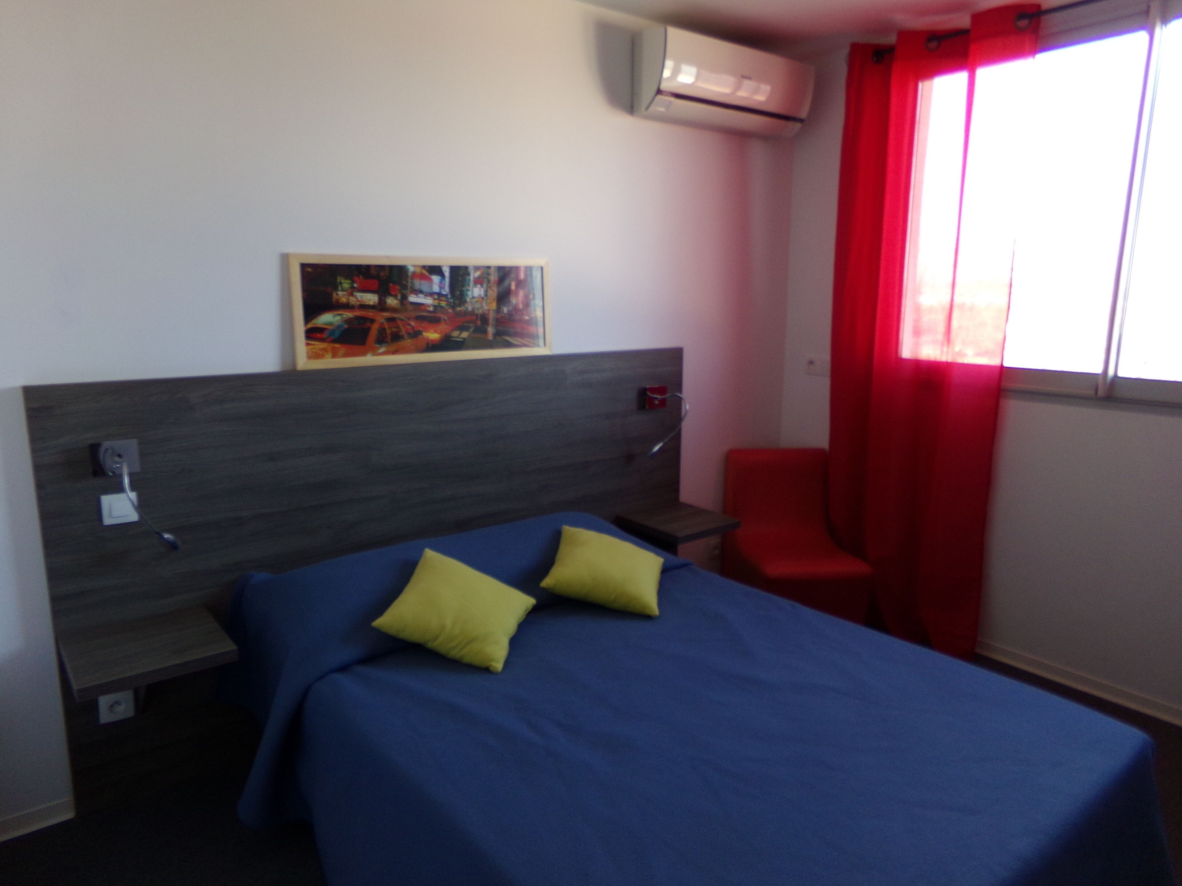 Una camera d'albergo con un letto, un comodino, una sedia rossa, un quadro appeso al muro e un condizionatore d'aria.