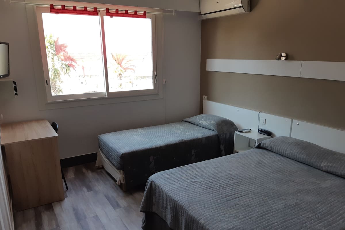Une chambre avec deux lits, un bureau en bois, une table de chevet et une fenêtre avec un rideau rouge.