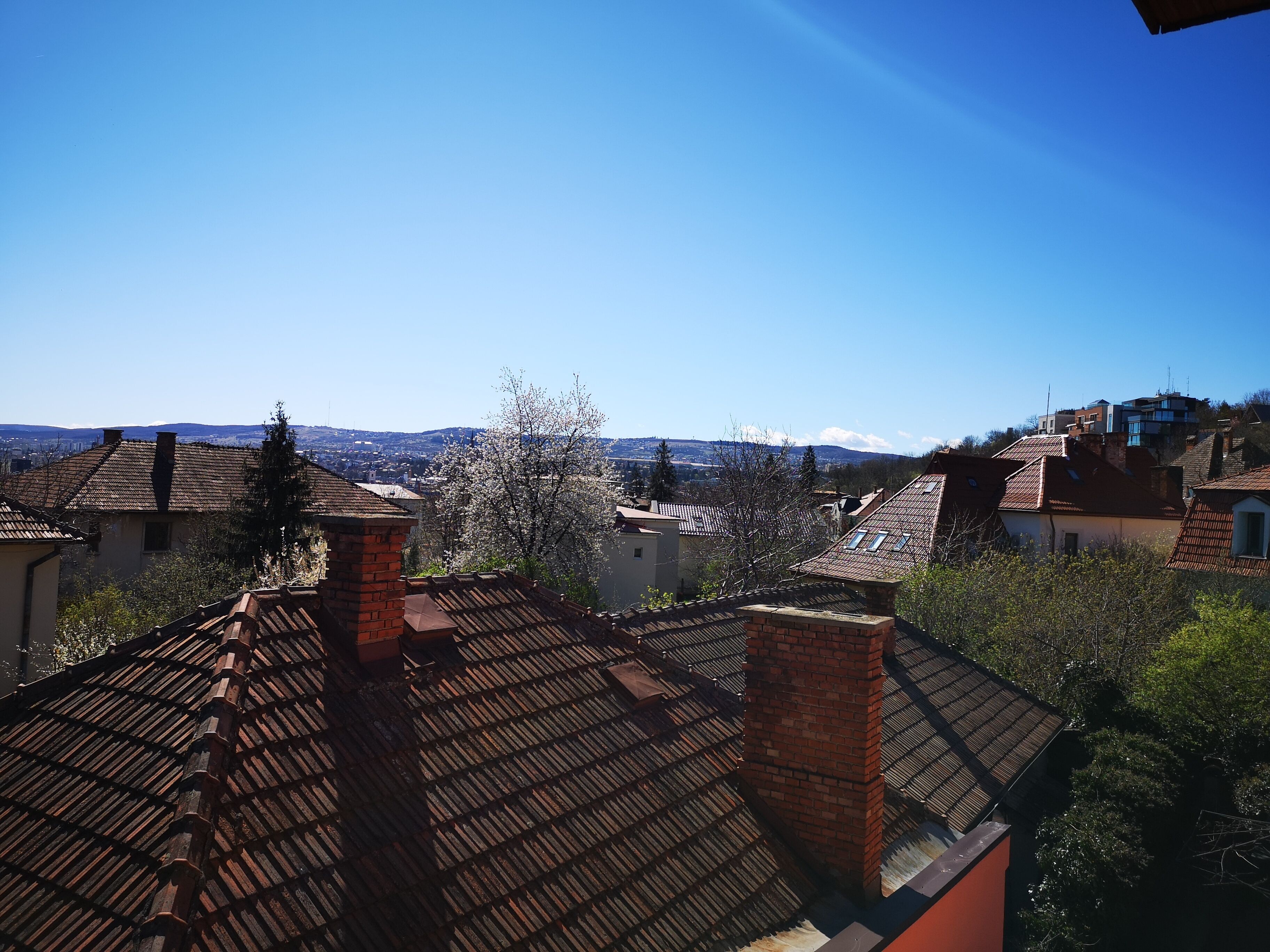 A residential area with terracotta-tiled roofs, brick chimneys, and a clear blue sky.
