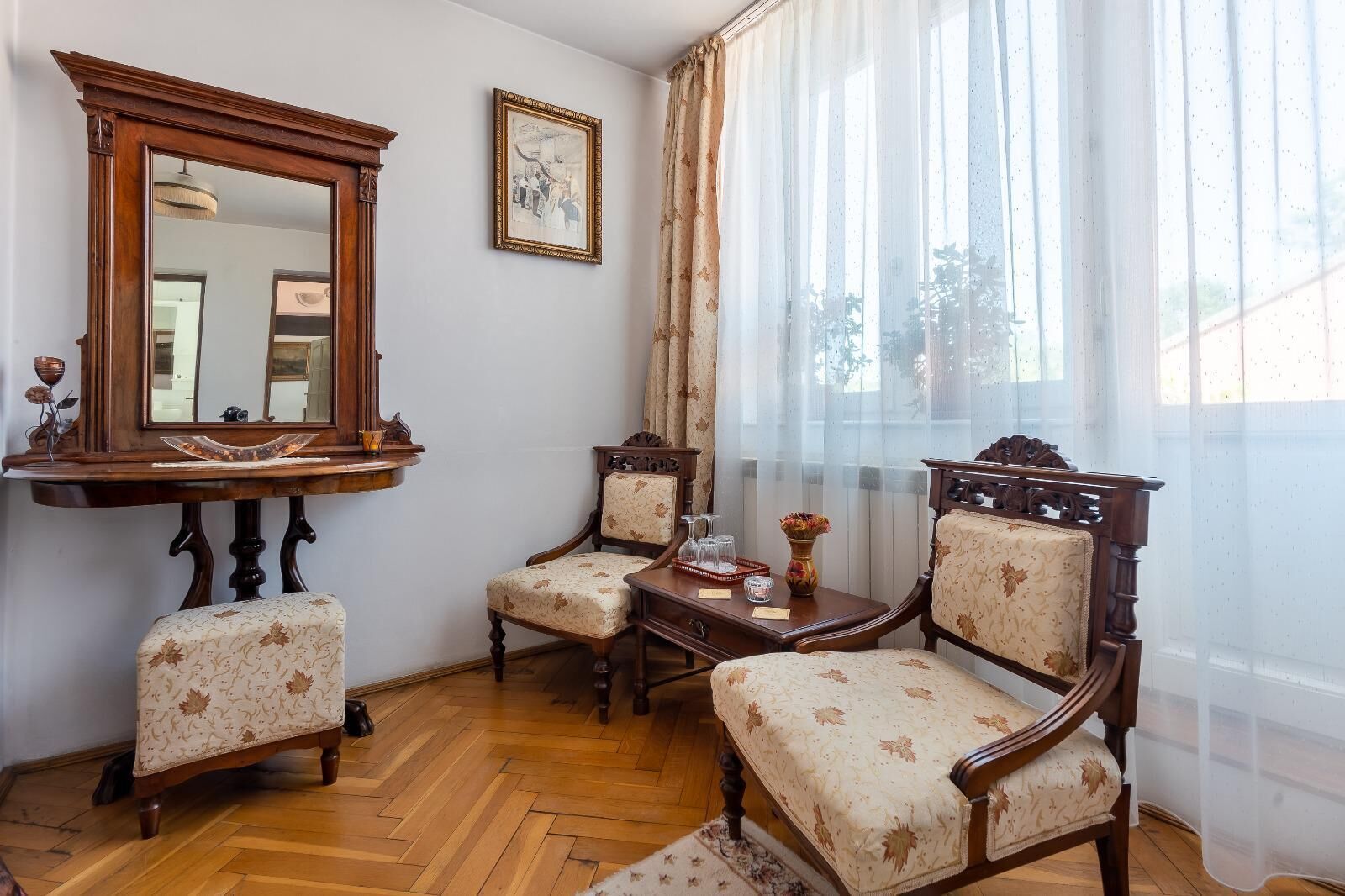 Habitació amb terra de fusta, un mirall gran, dues butaques tapissades, una taula petita amb un vas i cortines transparents.