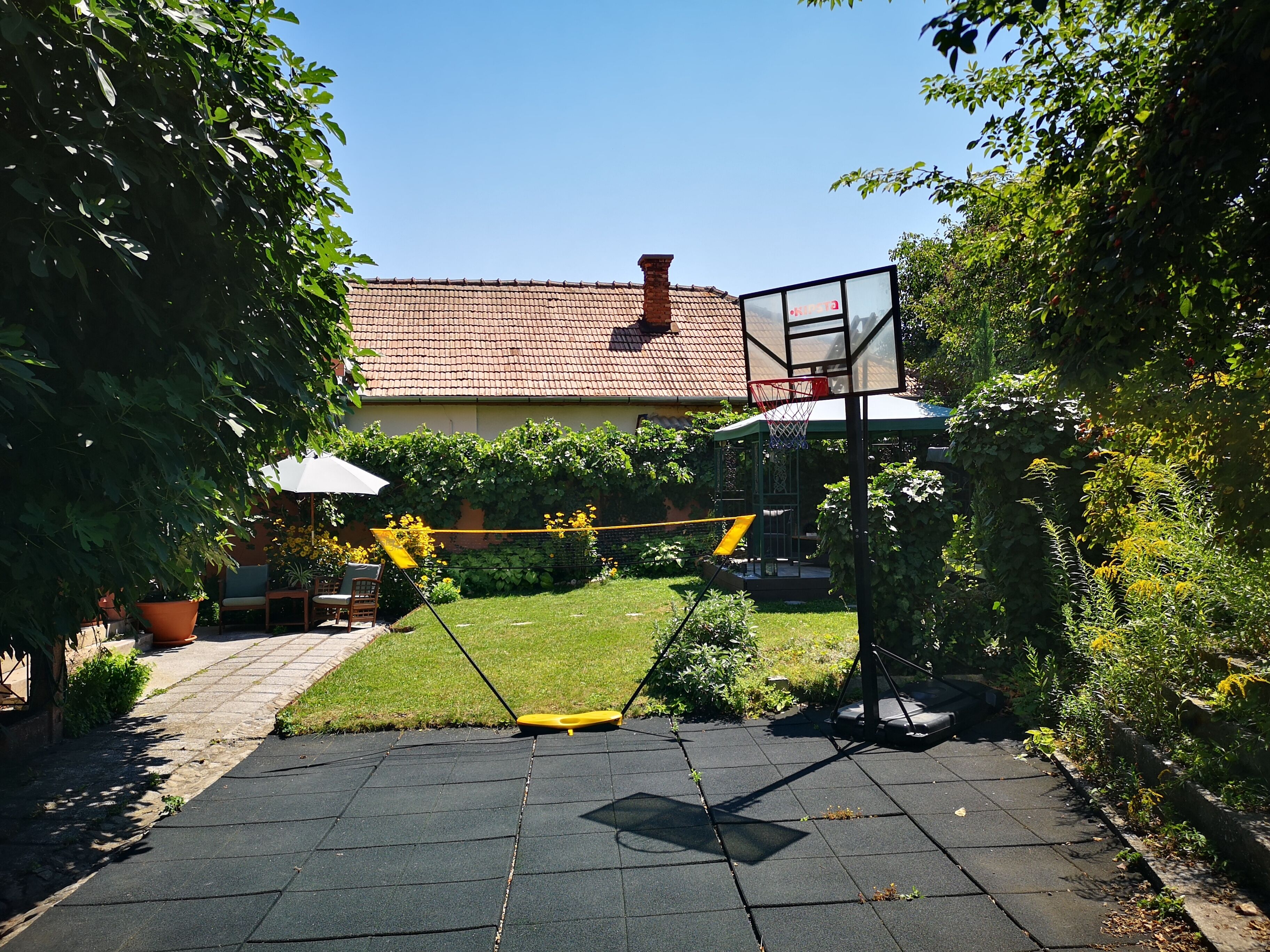 Un jardin avec un panier de basket, un espace patio avec des chaises et une table, et une maison avec un toit rouge en arrière-plan.