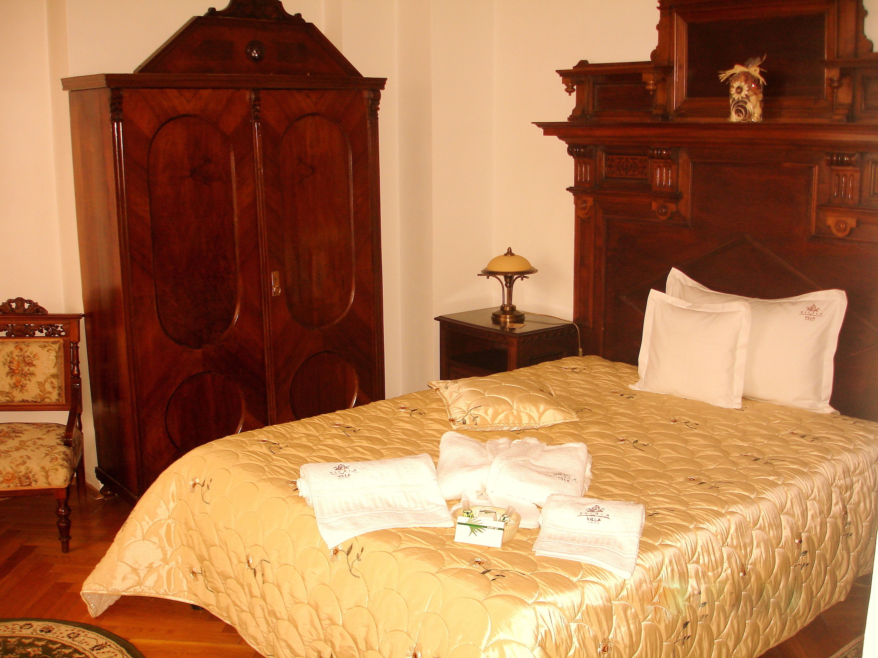 A hotel room with a large wooden wardrobe, a bedside table with a lamp, a bed with a quilt and towels, and a floral-patterned chair.