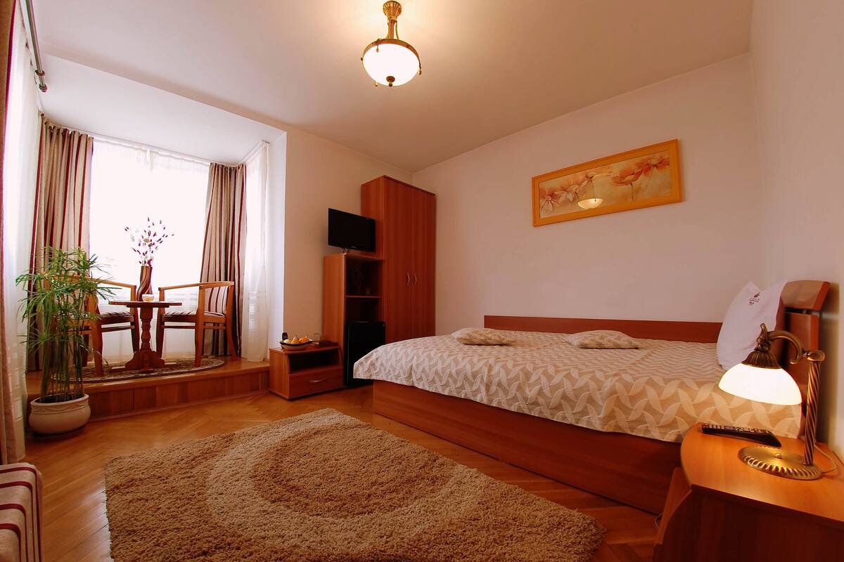 Un dormitorio con cama, mesita de noche con lámpara, televisión, armario, silla, mesa con planta y un cuadro en la pared.