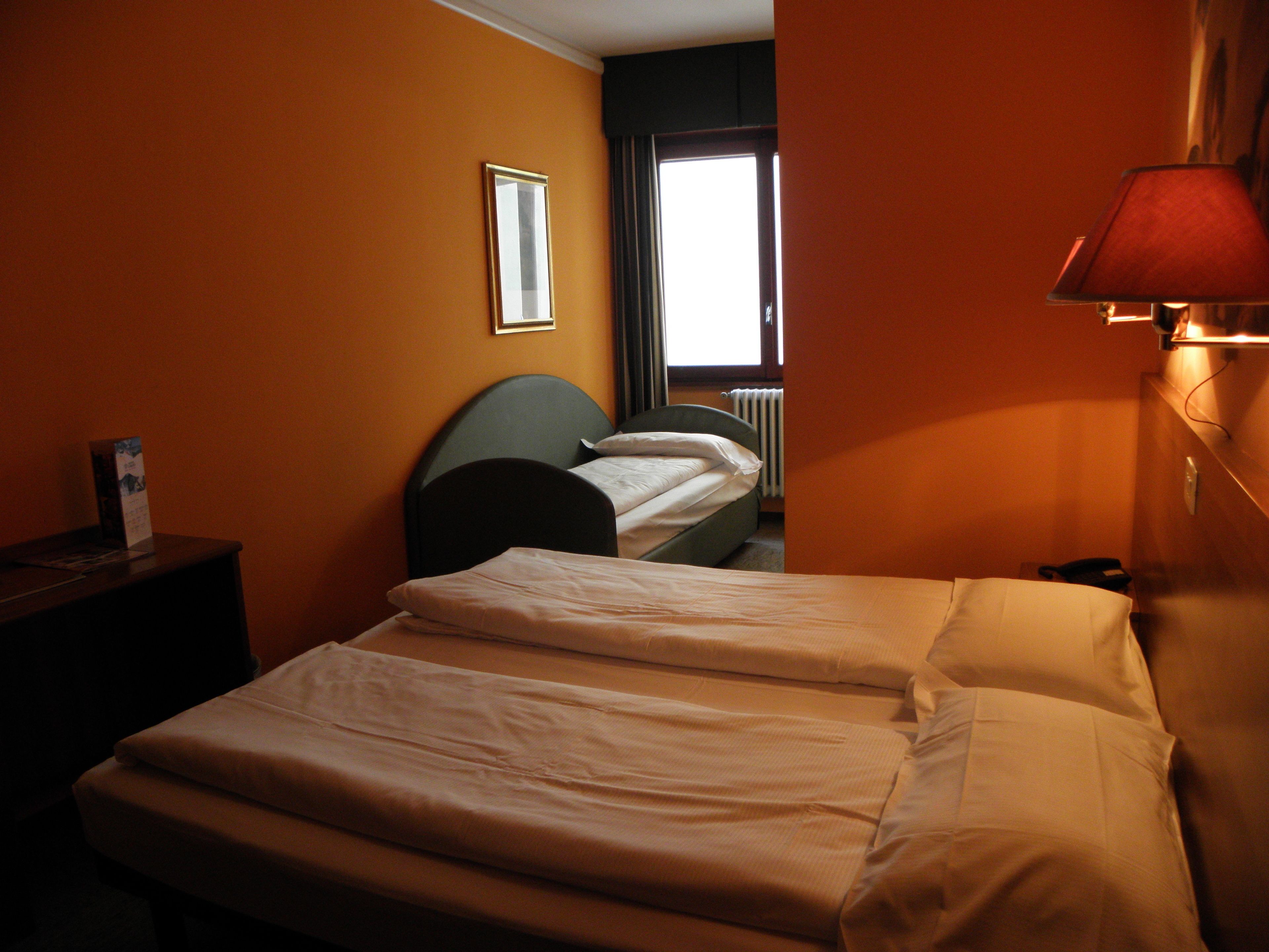 A hotel room with two beds, a nightstand, a lamp, and a window with curtains.