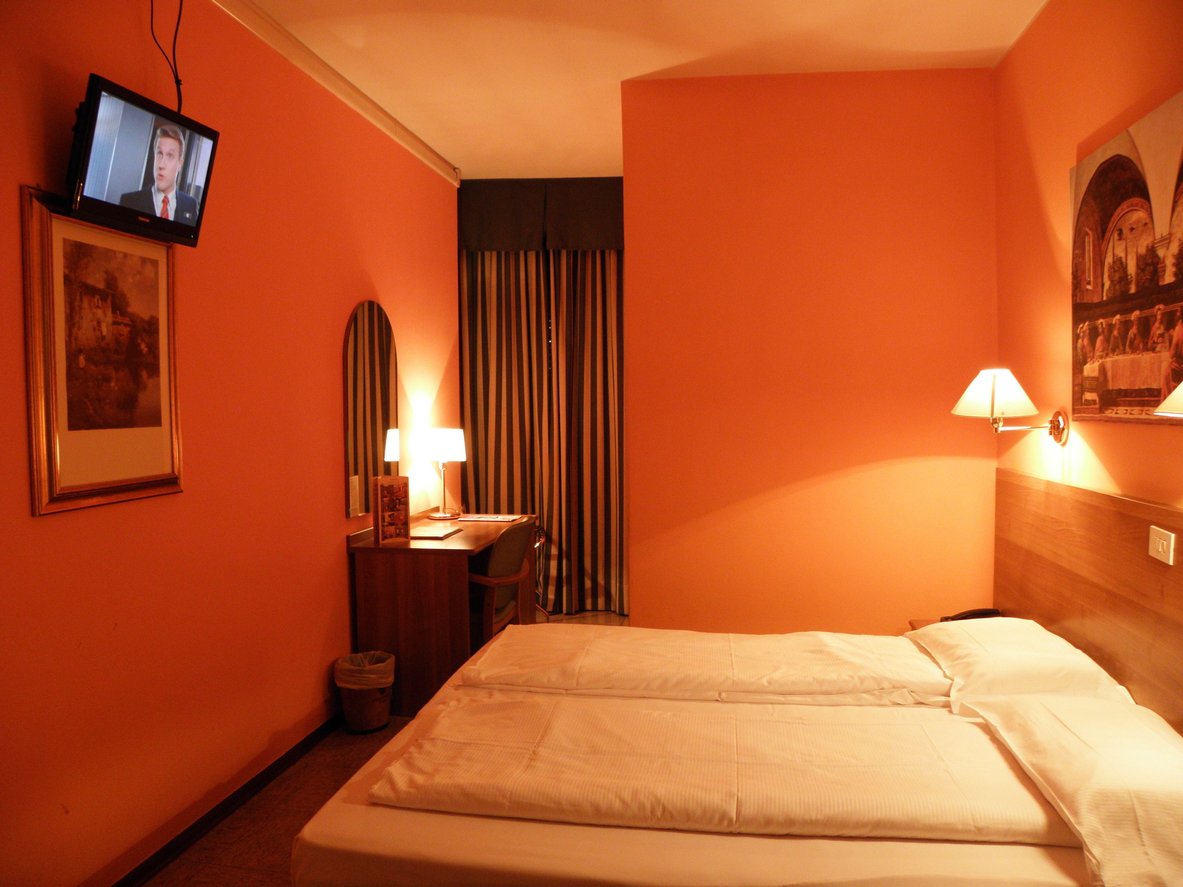 A hotel room with a bed, a desk with a lamp, a television on the wall, and a framed picture.