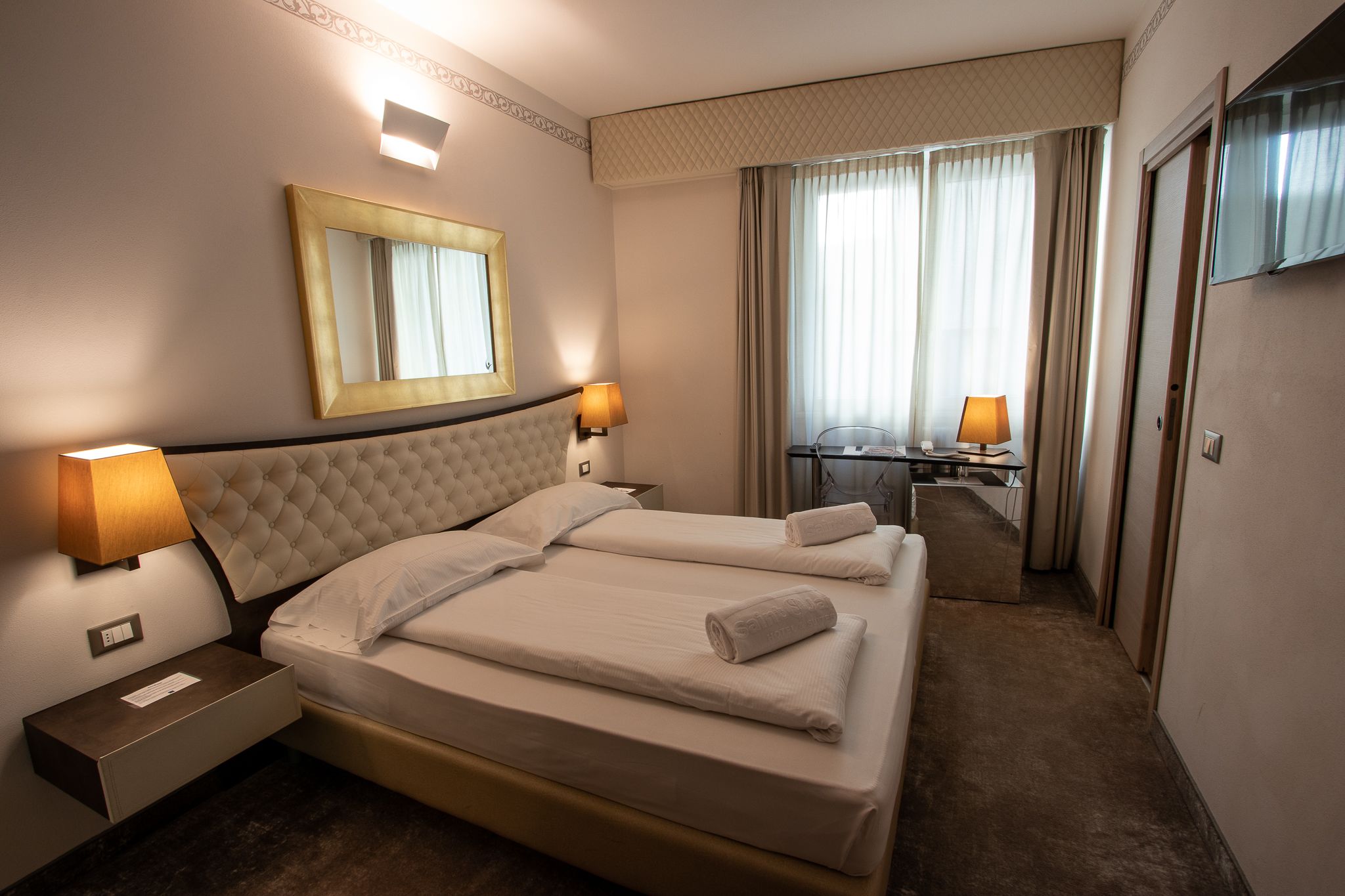 A hotel room with two beds, a large mirror, a desk with a chair, and a TV on the wall.