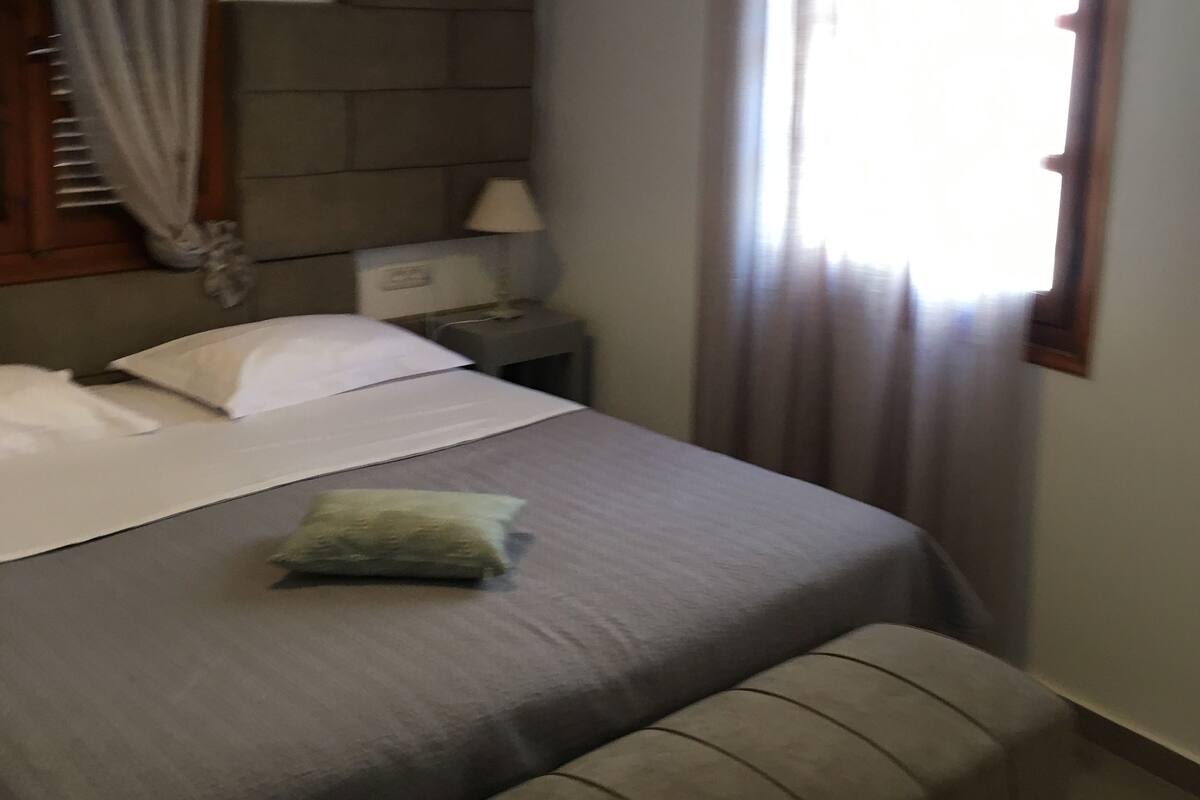 Une chambre à coucher avec un lit, une table de chevet, une lampe, un banc et une fenêtre avec des rideaux.