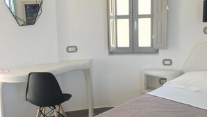A bedroom with a bed, a desk, a chair, a window, and a wall-mounted mirror. - Argonaftes (Santorini)