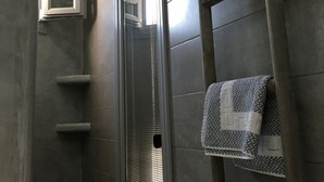 A bathroom with a shower, a window with blinds, a towel rack, and a towel on the rack. - Argonaftes (Santorini)