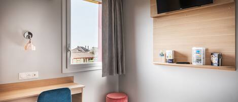 A hotel room with a bed, a desk, a chair, a TV, a window with curtains, and a shelf with items.