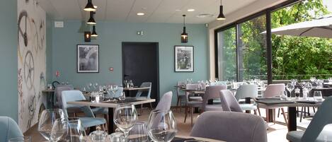 A restaurant with tables set for dining, a large window with a view of greenery, and hanging pendant lights.