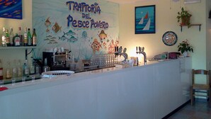 Un coin bar orné d’une fresque représentant des poissons et sur lequel on peut lire : « Trattoria del Pesce Povero ». On y trouve des bouteilles et des verres sur le comptoir, un ventilateur au plafond et une chaise dans un coin.