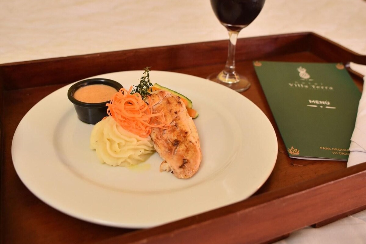 Un plateau avec une assiette de nourriture, un verre de vin rouge et un menu, le tout posé sur un lit doté d’une tête de lit en bois.