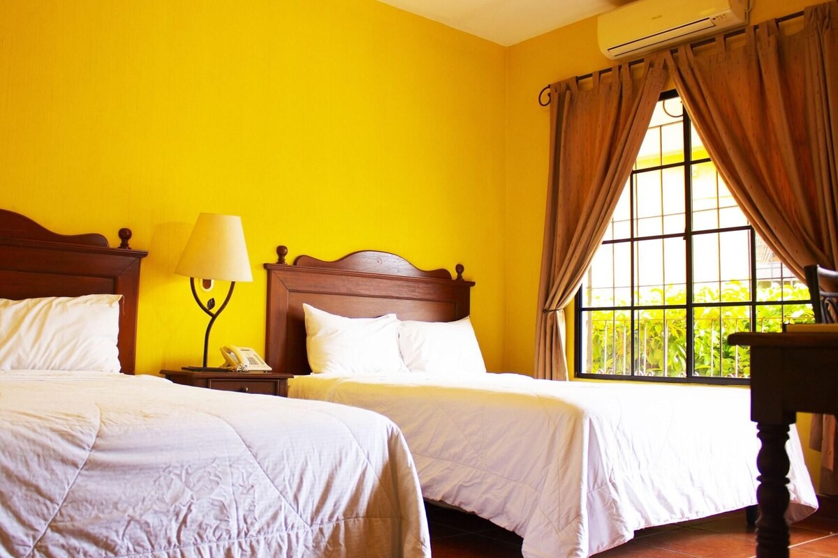 A hotel room with two beds, a nightstand with a lamp, and a window with curtains.