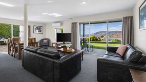 A spacious living room with a large black leather sofa, a dining area with wooden furniture, a flat-screen TV, and a balcony with a scenic view.