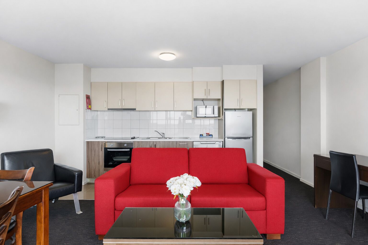 A modern living room with a red sofa, a glass coffee table, and a kitchen area with white cabinets and appliances.