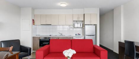 A modern living room with a red sofa, a glass coffee table, and a kitchen area with white cabinets and appliances.