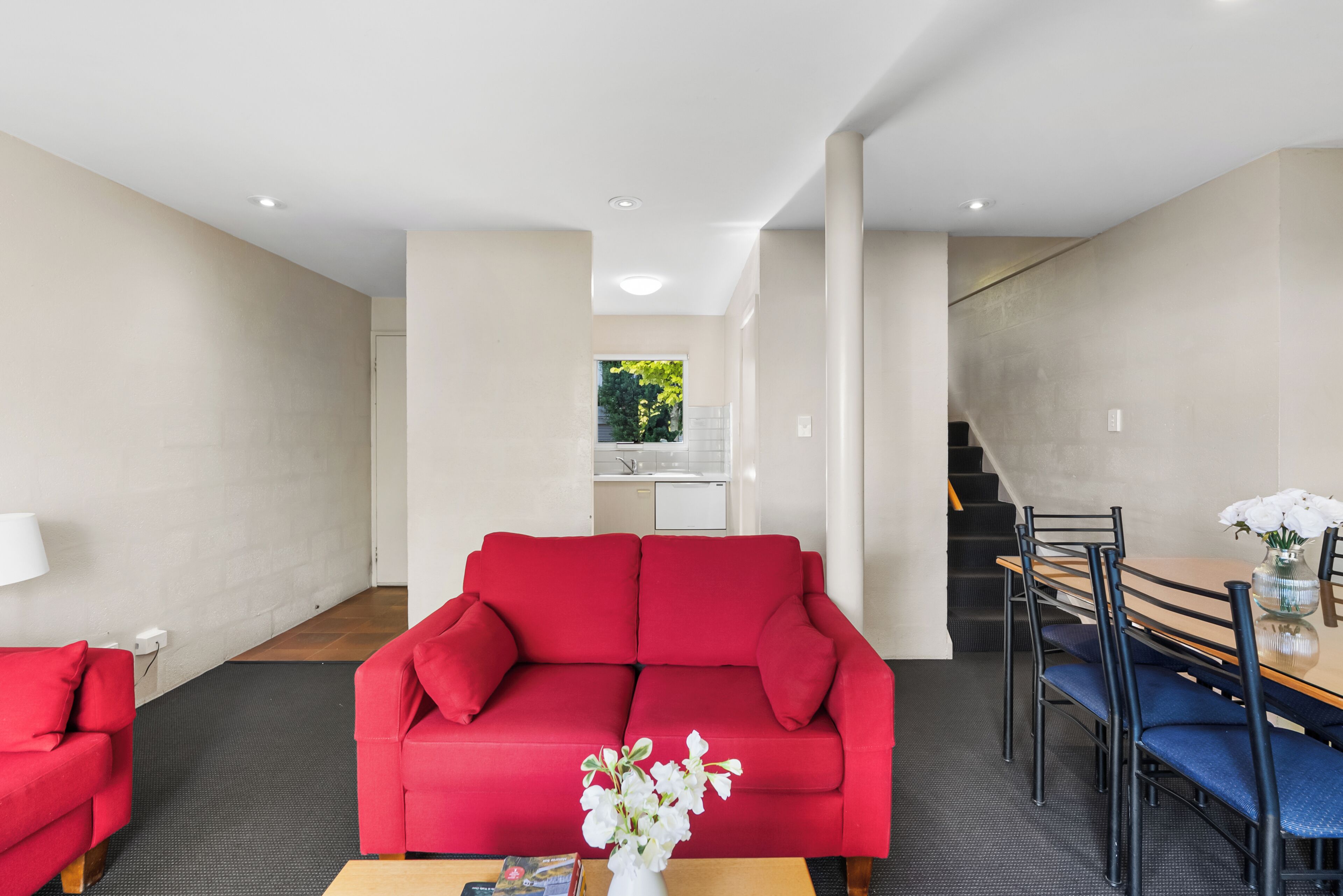 A modern living room with a red sofa, a dining table with chairs, a small kitchen area, and a staircase.