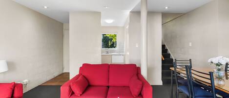 A modern living room with a red sofa, a dining table with chairs, a small kitchen area, and a staircase.