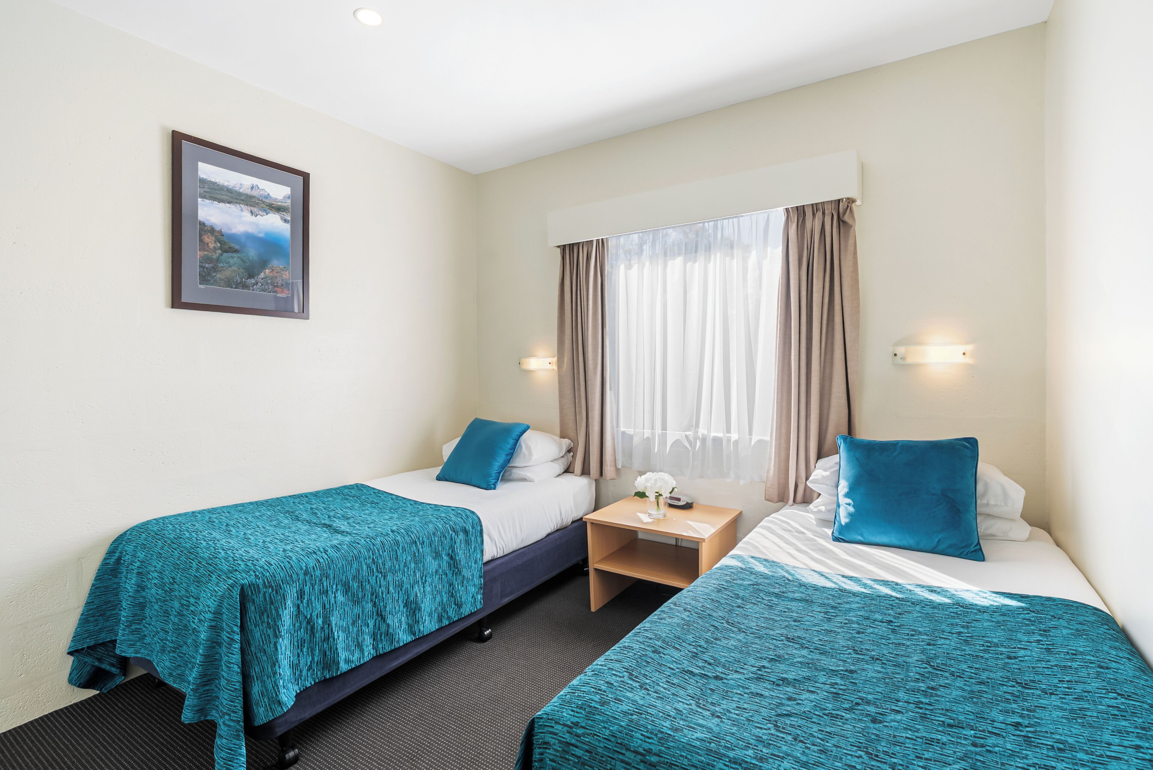 A hotel room with two beds, a nightstand, a framed picture on the wall, and a window with curtains.
