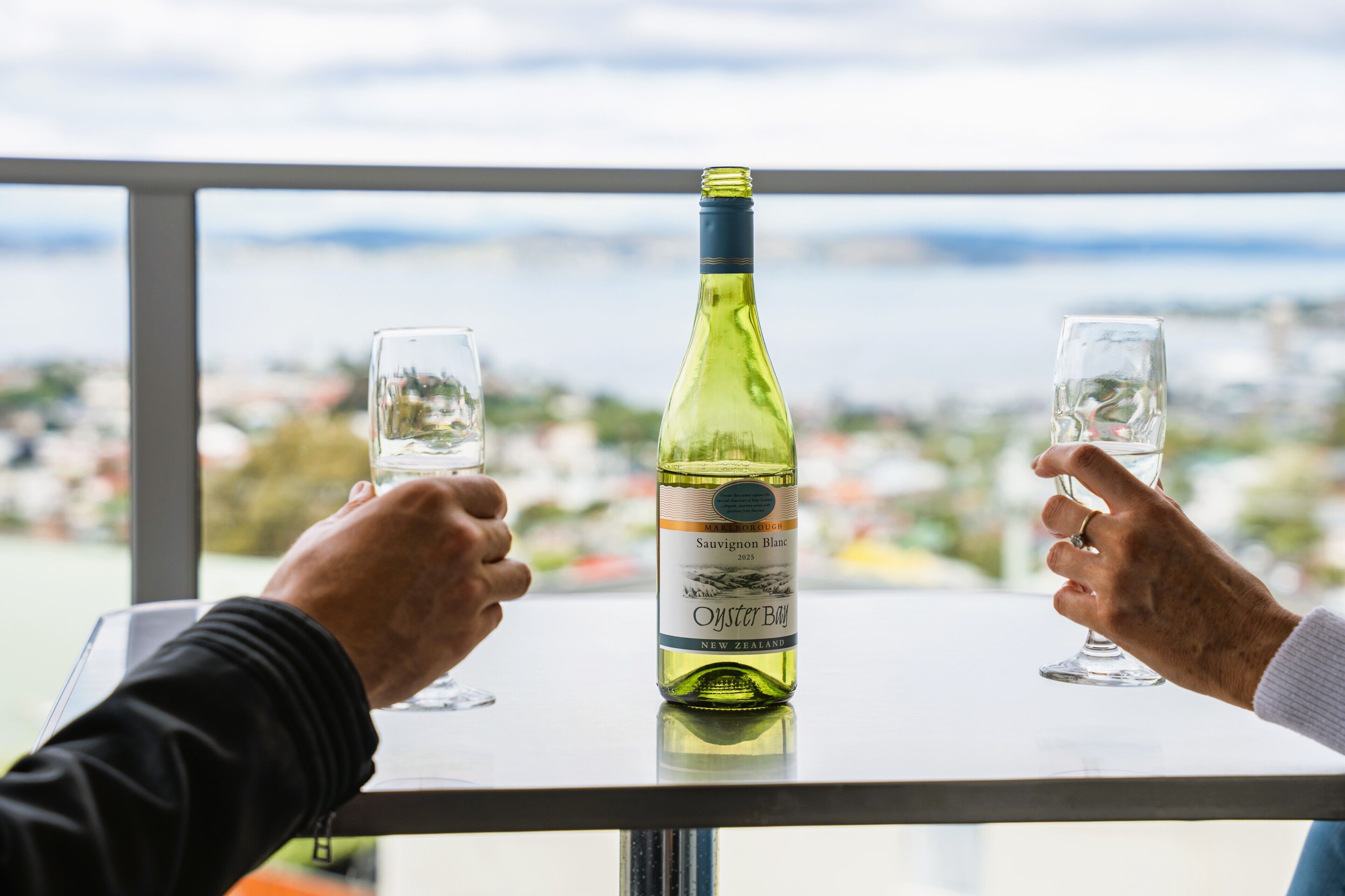 Two hands holding wine glasses on a table with a bottle of Sauvignon Blanc in the center.