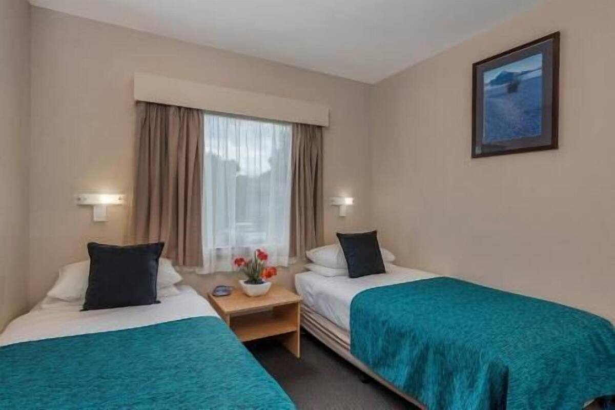 A hotel room with two beds, a nightstand, a window with curtains, and a painting on the wall.