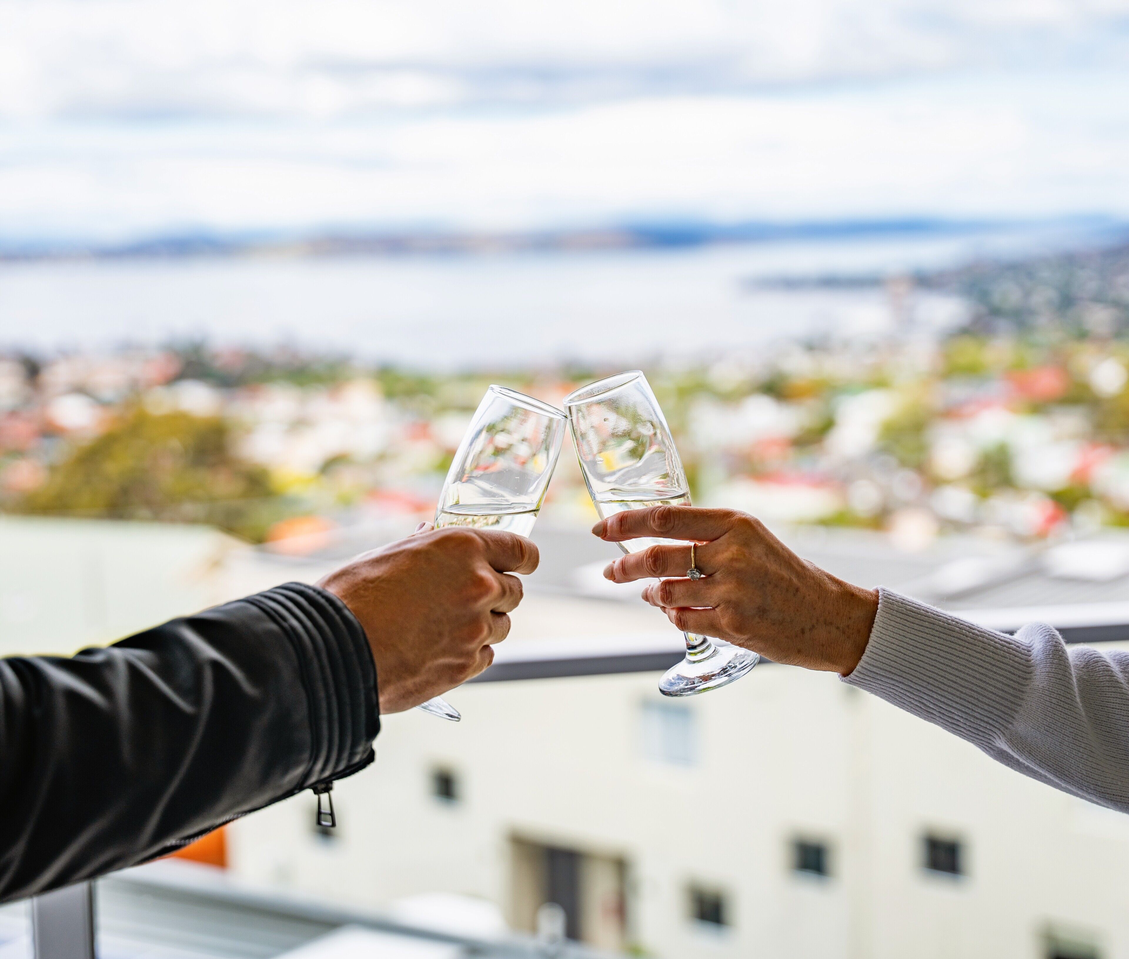 Dos manos sosteniendo copas de vino, con vistas a un paisaje urbano que incluye edificios y una masa de agua.