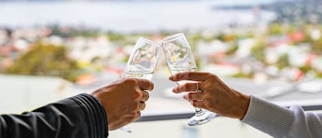 Two hands holding wine glasses, overlooking a cityscape with buildings and a body of water.