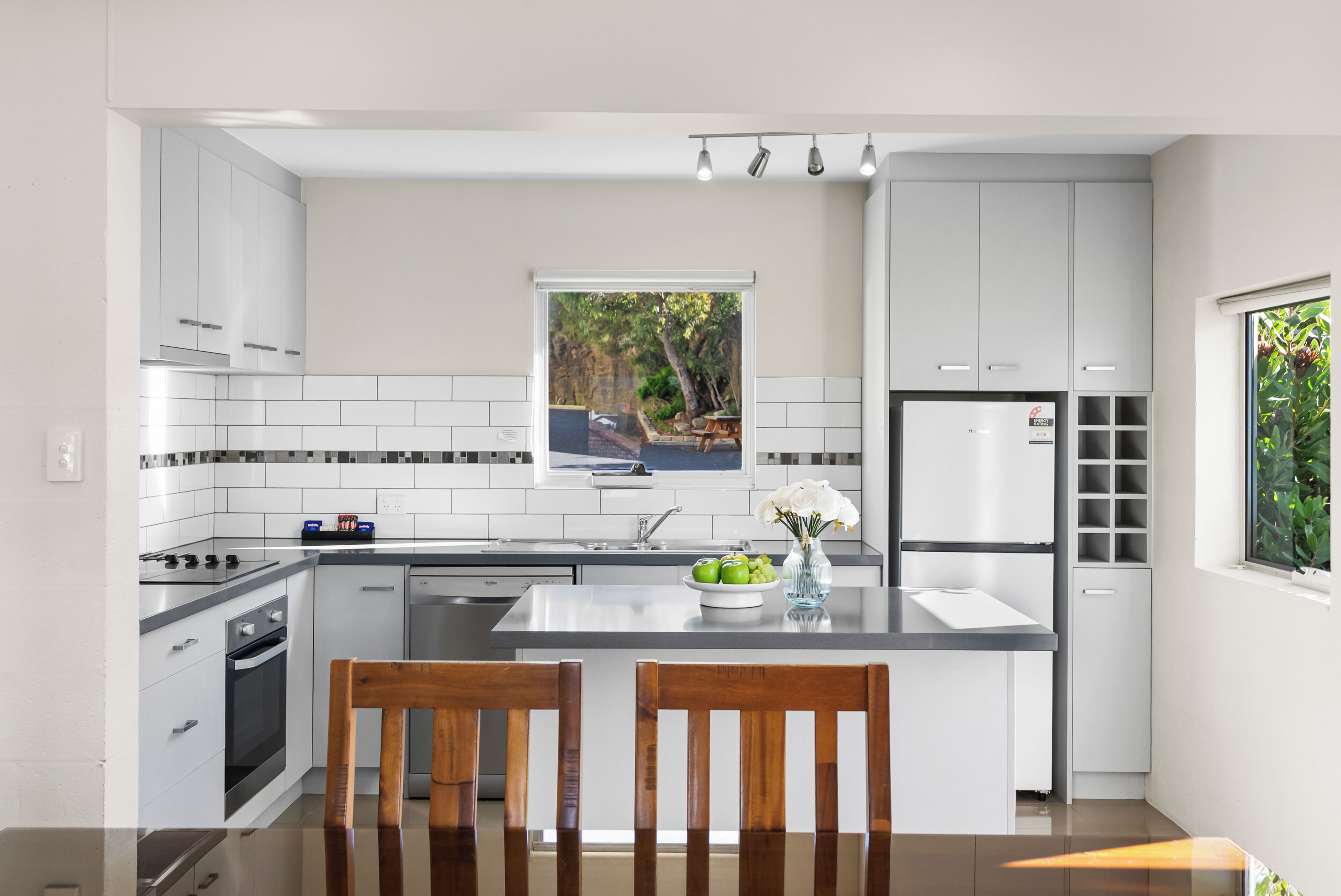 Una cucina moderna con mobili bianchi, un'isola centrale, un forno integrato e una finestra con vista su un giardino.