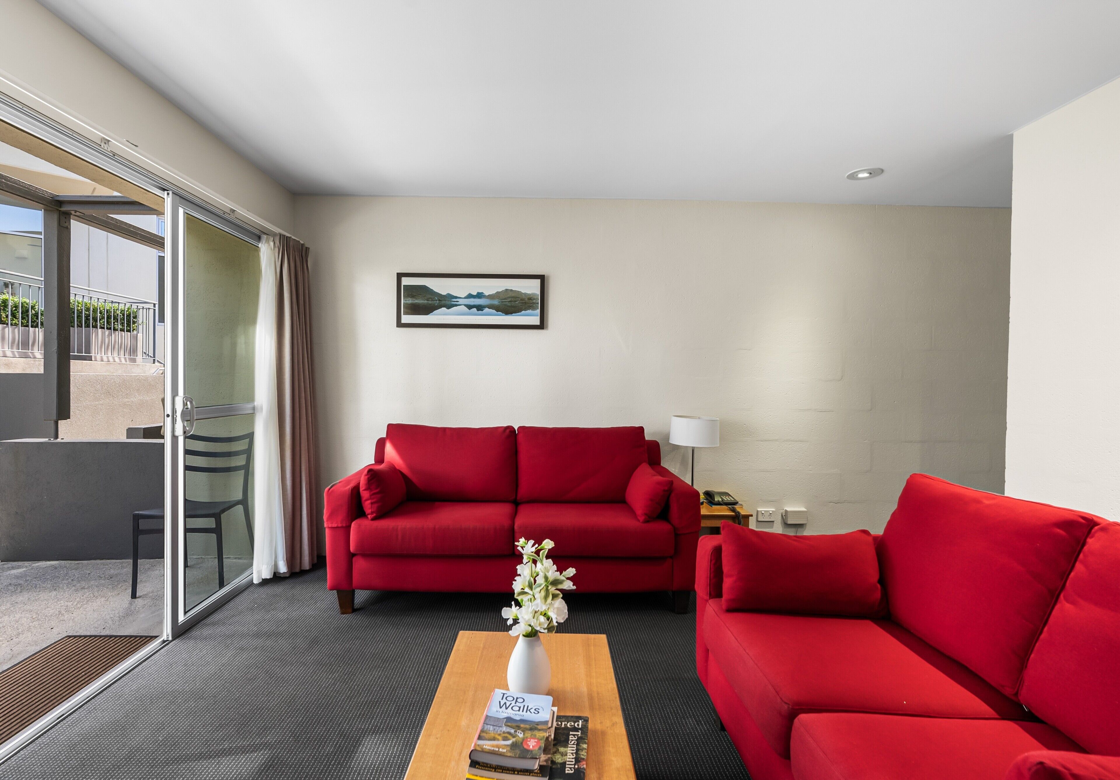 A modern hotel room with a red sofa, a wooden coffee table with books, a glass door leading to a balcony, and a painting on the wall.