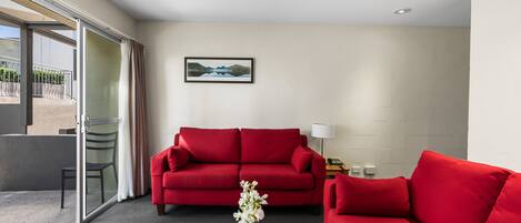A modern hotel room with a red sofa, a wooden coffee table with books, a glass door leading to a balcony, and a painting on the wall.