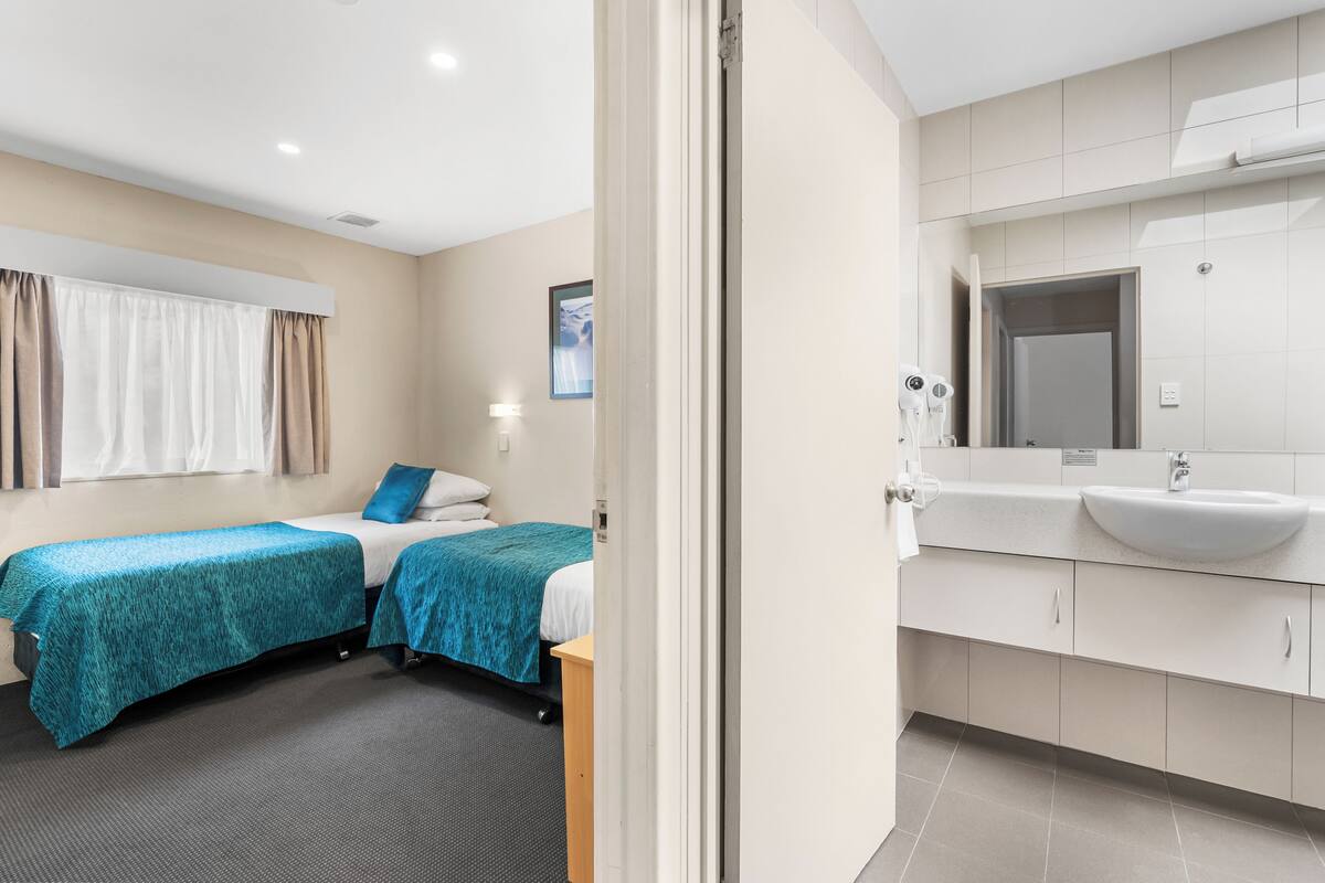 A hotel room with two beds, a bathroom with a sink and mirror, and a window with curtains.