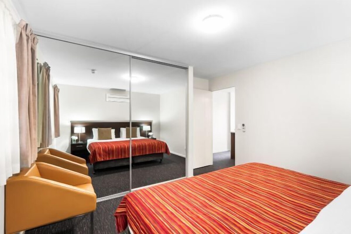 A hotel room with a mirrored closet, a bed with a striped bedspread, a chair, and a bedside table.