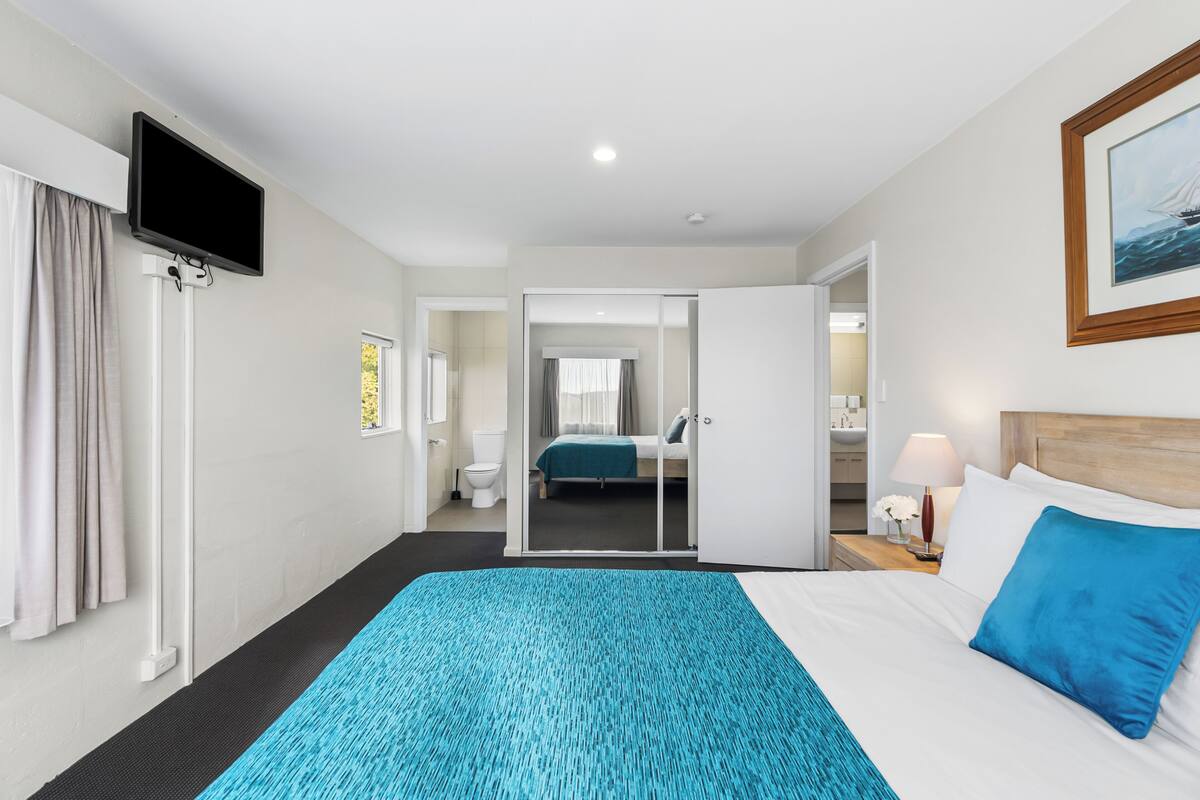 A hotel room with a bed, a TV mounted on the wall, a bathroom with a toilet, and a mirror.