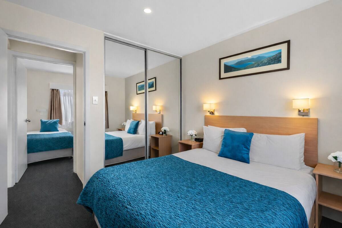 A hotel room with a large bed, a mirror, a nightstand, a wall-mounted lamp, and a framed picture.