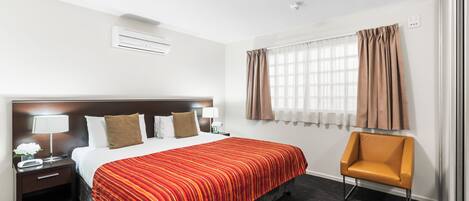 Una habitación de hotel con una cama, una silla, una mesita de noche, una lámpara y una ventana con cortinas.